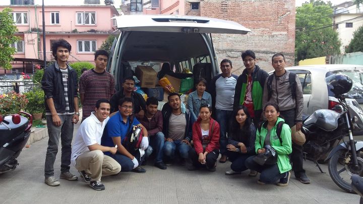 Fifteen volunteers stand and crouch in two rows behind a van with its tailgate raised, the load space filled with cardboard boxes and sacks of relief supplies.