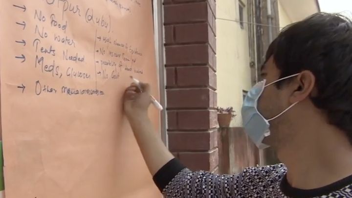 A masked volunteer writes on a large sheet of paper taped to a wall, listing needs in a column: no food, no water, tents needed, meds, glucose.