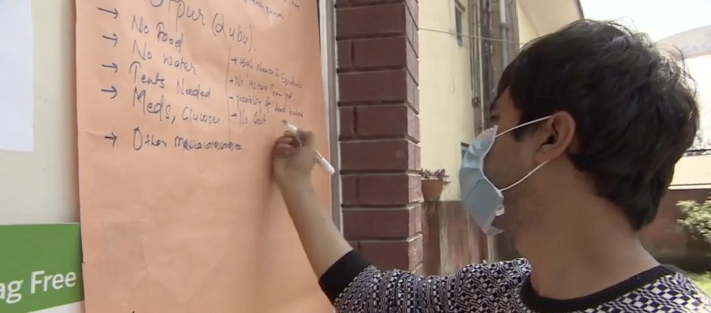 A masked volunteer writes on a large sheet of paper taped to a wall, listing needs in a column: no food, no water, tents needed, meds, glucose.
