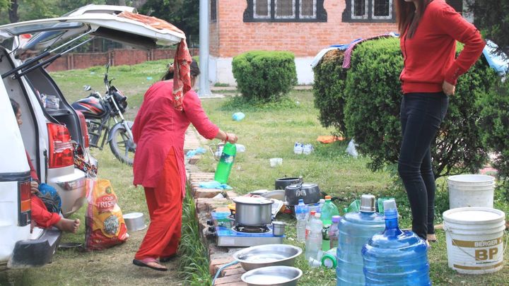 A woman in a red kurta pours water from a bottle beside a car with its boot open, next to cooking pots on a portable gas stove, steel bowls and large blue water jars set out on a lawn.