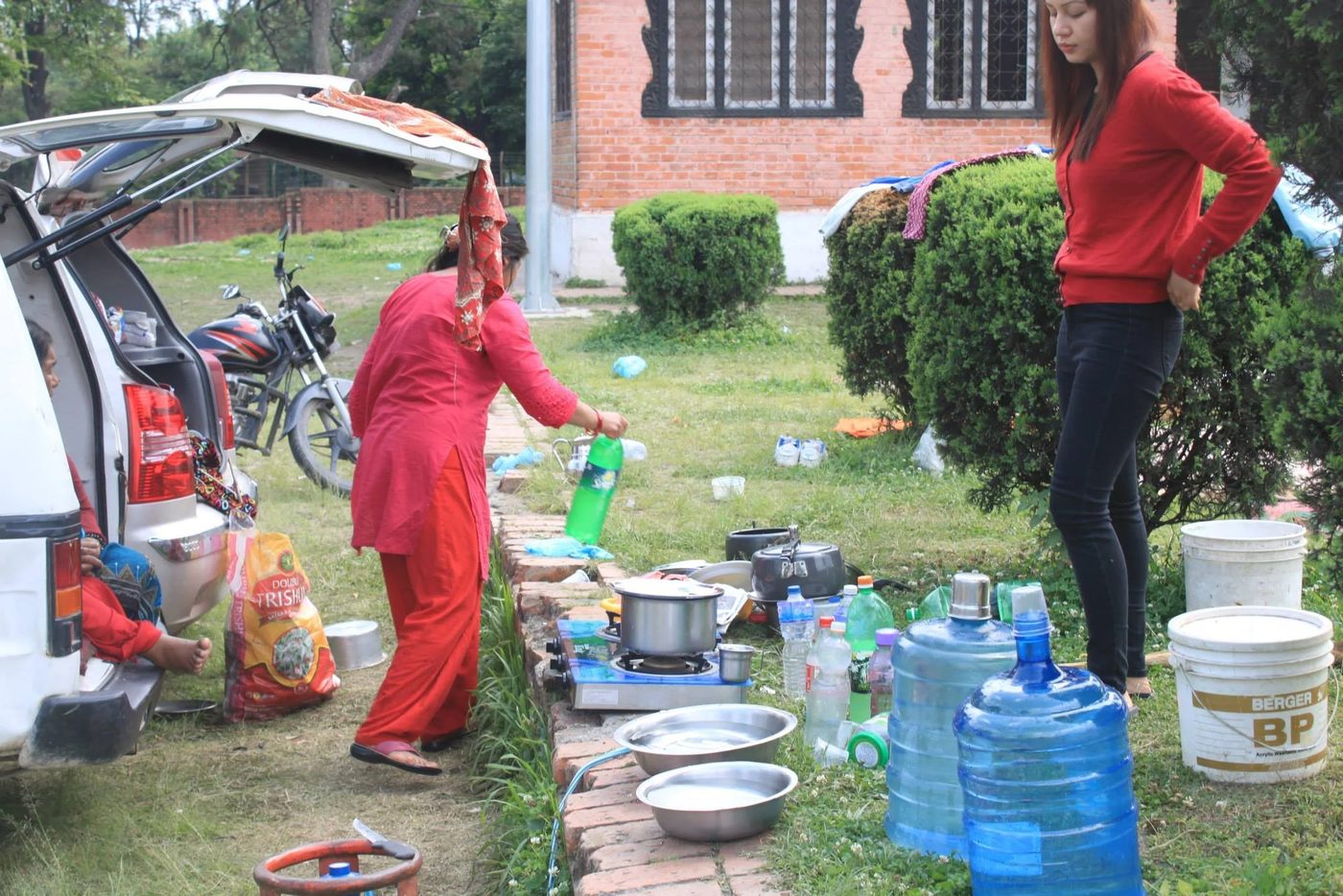 A woman in a red kurta pours water from a bottle beside a car with its boot open, next to cooking pots on a portable gas stove, steel bowls and large blue water jars set out on a lawn.