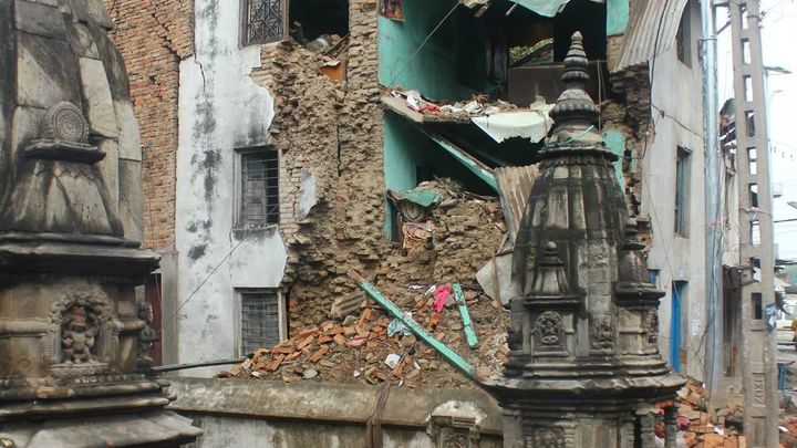 A three storey house with its front wall torn away, exposing the rooms inside, with brick rubble heaped at its base beside carved stone shrines on a wet street.