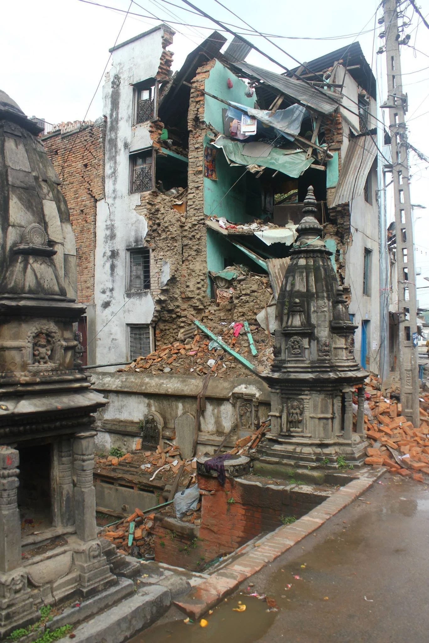 A three storey house with its front wall torn away, exposing the rooms inside, with brick rubble heaped at its base beside carved stone shrines on a wet street.