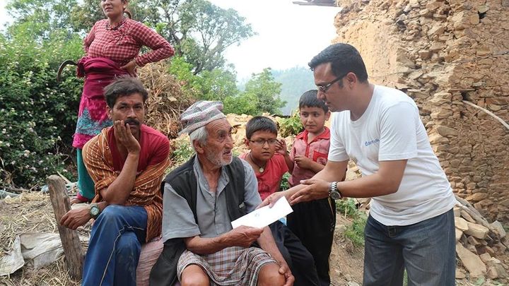 A volunteer in a white Deerwalk t-shirt hands an envelope to an elderly man seated on a sack, watched by a family of two women and two boys beside a collapsed stone wall.