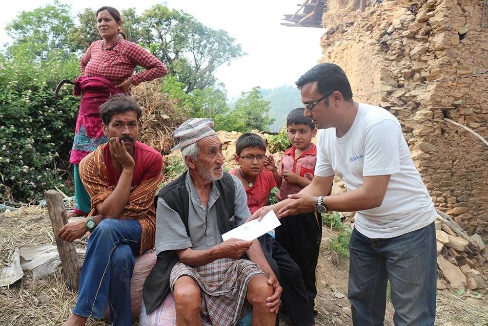 A volunteer in a white Deerwalk t-shirt hands an envelope to an elderly man seated on a sack, watched by a family of two women and two boys beside a collapsed stone wall.