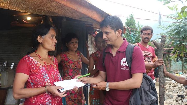 A volunteer in a Deerwalk polo shirt holds out a form and a pen to a woman outside a bamboo and thatch shelter, watched by two girls and a man, with rubble at the right.