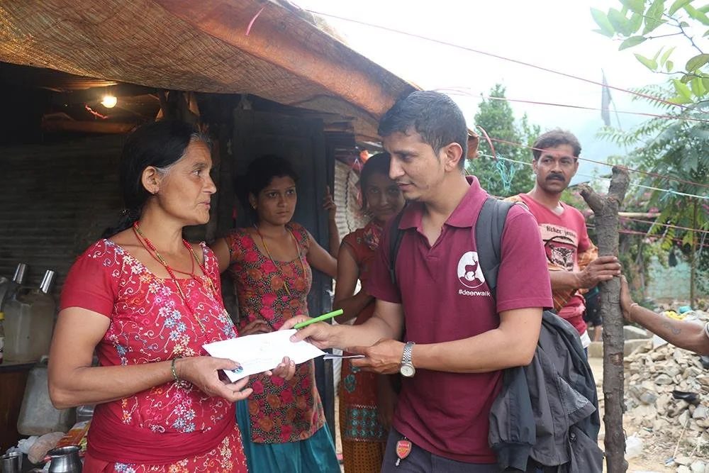 A volunteer in a Deerwalk polo shirt holds out a form and a pen to a woman outside a bamboo and thatch shelter, watched by two girls and a man, with rubble at the right.