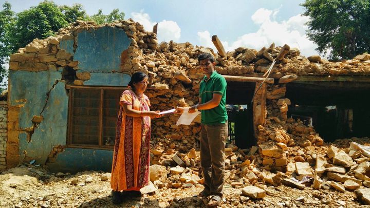 A woman in an orange dress takes an envelope from a volunteer in a green shirt as they stand on the rubble of a stone house whose walls have collapsed.
