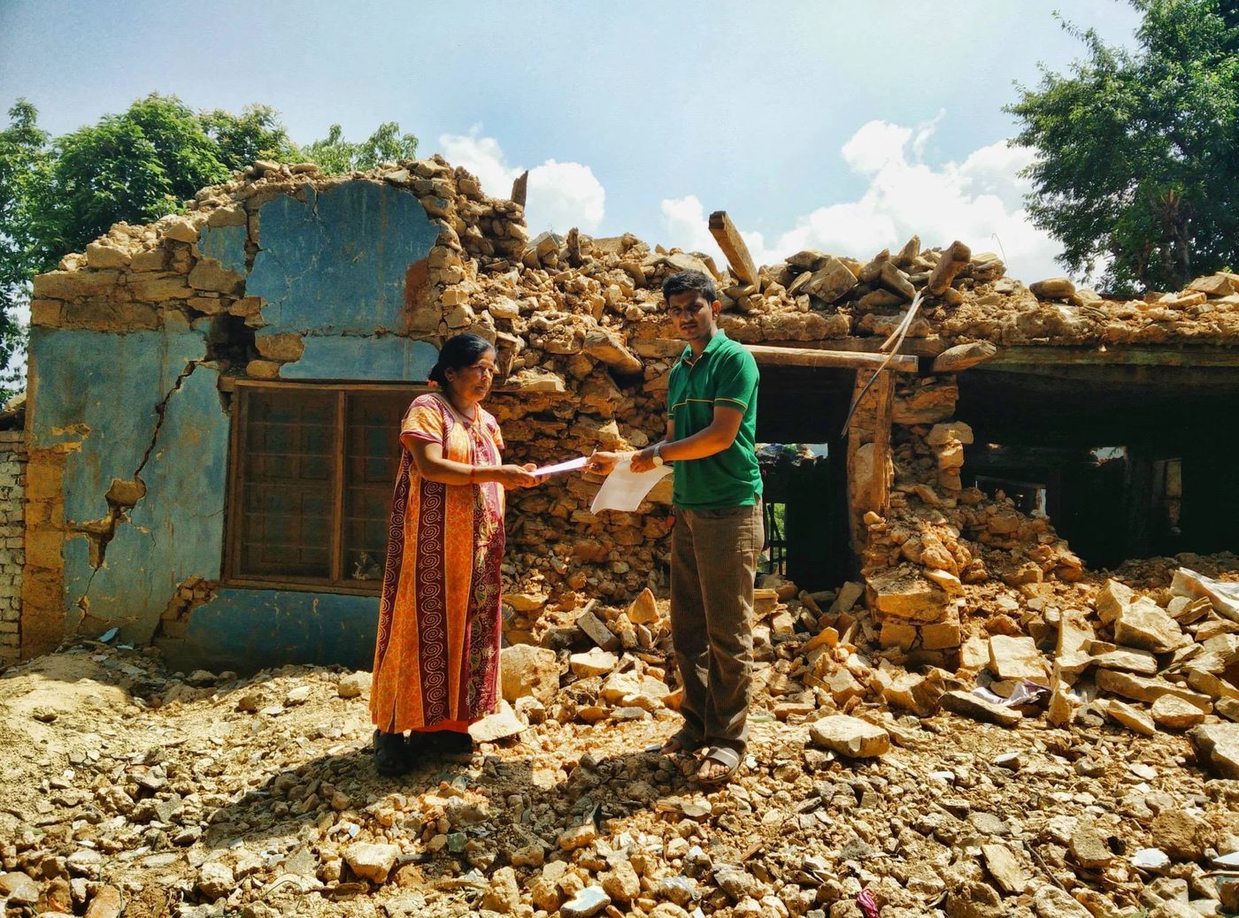 A woman in an orange dress takes an envelope from a volunteer in a green shirt as they stand on the rubble of a stone house whose walls have collapsed.