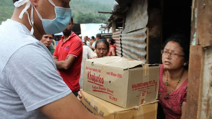 A volunteer in a face mask lifts two stacked cardboard cartons of supplies towards a woman standing in the doorway of a corrugated iron shelter, with people waiting behind him.