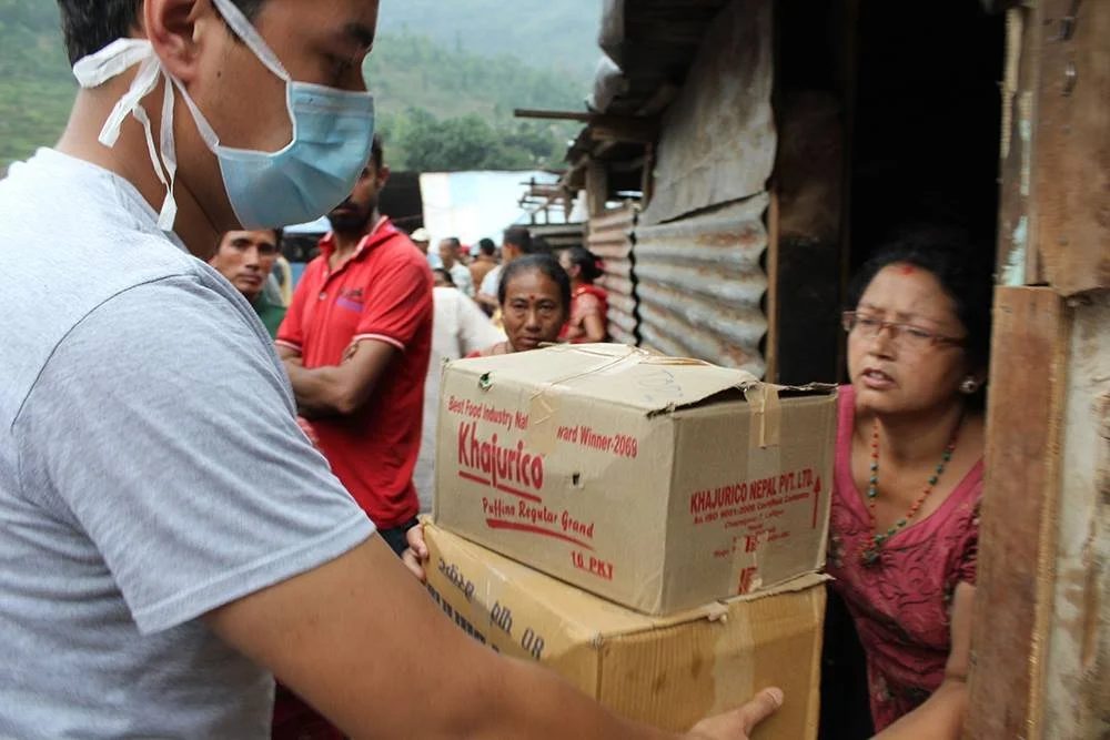 A volunteer in a face mask lifts two stacked cardboard cartons of supplies towards a woman standing in the doorway of a corrugated iron shelter, with people waiting behind him.