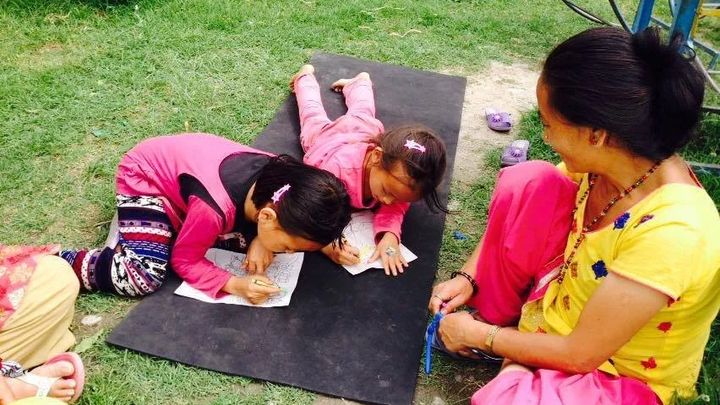 Two girls in pink lie on a black mat on a lawn colouring in outlined drawings, while a woman in a yellow blouse sits cross legged beside them.
