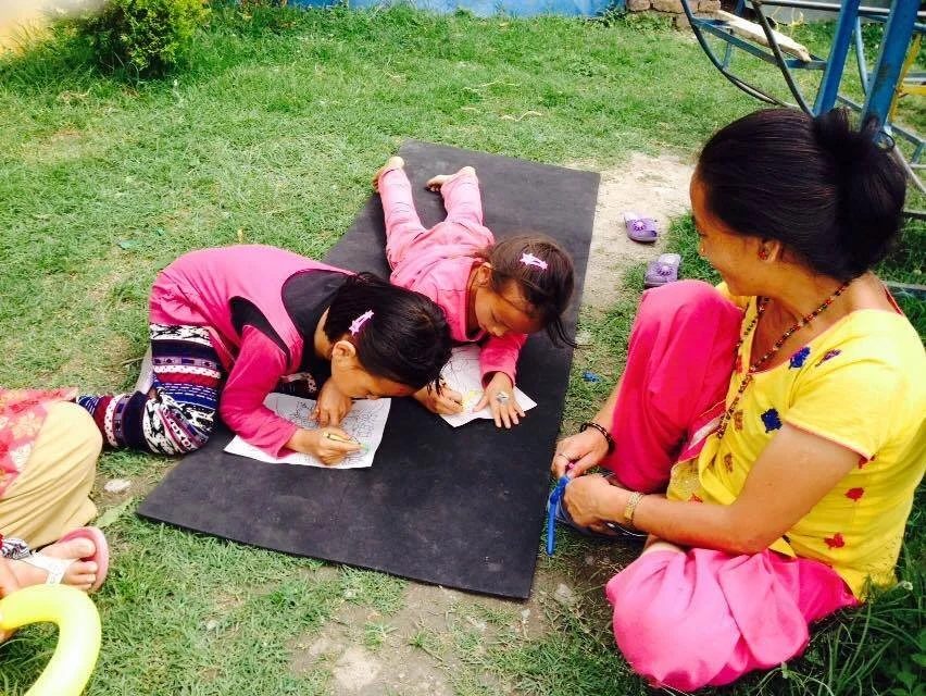 Two girls in pink lie on a black mat on a lawn colouring in outlined drawings, while a woman in a yellow blouse sits cross legged beside them.