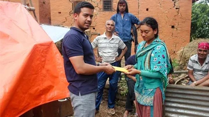 A volunteer hands folded banknotes to a young woman in a green kurta outside a cracked brick house, watched by three men standing beside a tent and a sheet of corrugated iron.