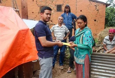 A volunteer hands folded banknotes to a young woman in a green kurta outside a cracked brick house, watched by three men standing beside a tent and a sheet of corrugated iron.