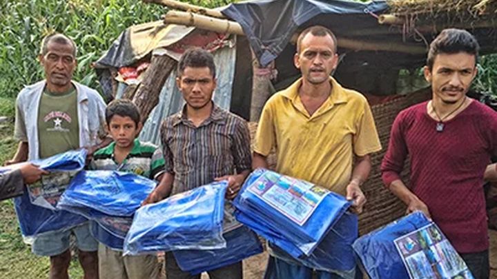 Five men and a boy stand in a row in front of a makeshift shelter, each holding a packaged blue tarpaulin sheet, with maize plants growing behind them.