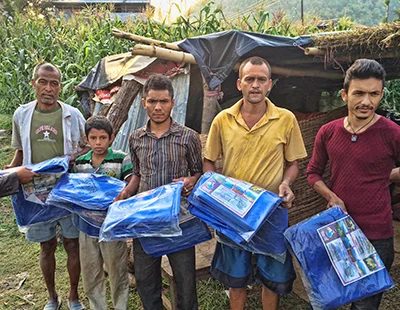 Five men and a boy stand in a row in front of a makeshift shelter, each holding a packaged blue tarpaulin sheet, with maize plants growing behind them.