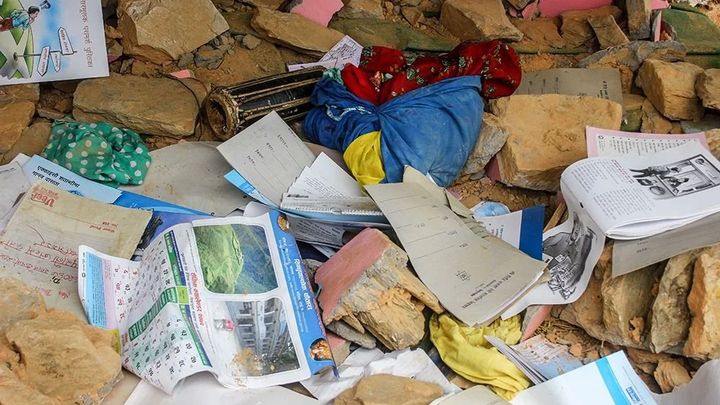School exercise books, a wall calendar and children's clothes lie scattered and open among stone rubble.