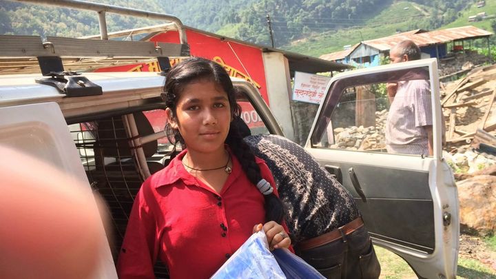 A girl in a red kurta stands beside a pickup truck holding a folded blue tarpaulin sheet, with a man at the open truck door and a green hillside behind.