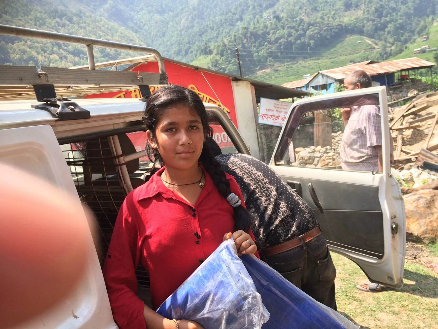 A girl in a red kurta stands beside a pickup truck holding a folded blue tarpaulin sheet, with a man at the open truck door and a green hillside behind.