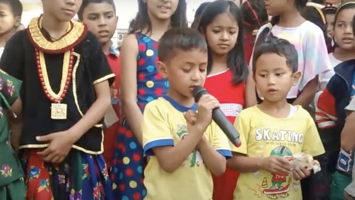 A young boy sings into a handheld microphone in a crowd of children in bright clothes, some in traditional dress, watching him closely.