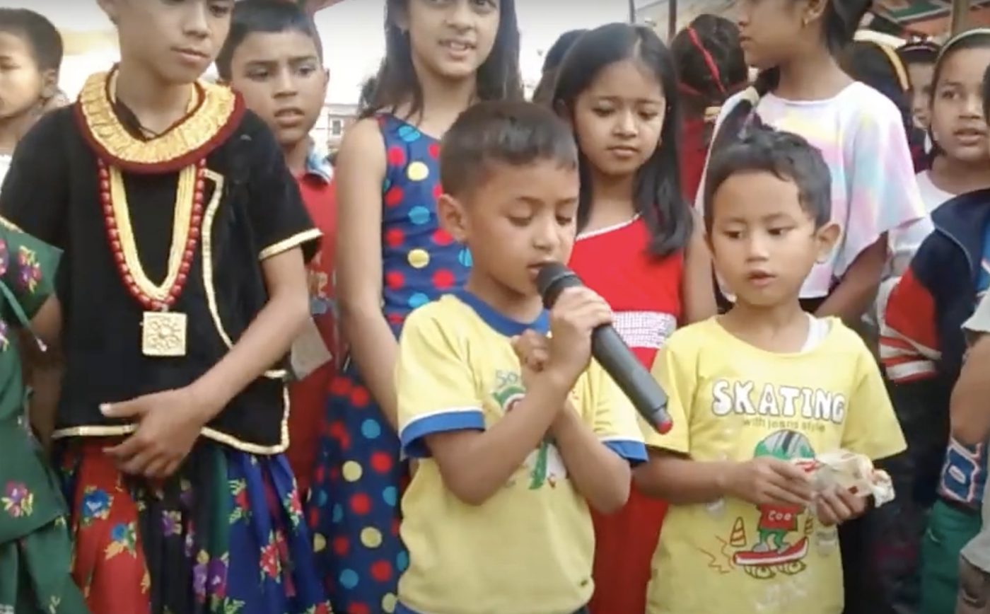 A young boy sings into a handheld microphone in a crowd of children in bright clothes, some in traditional dress, watching him closely.
