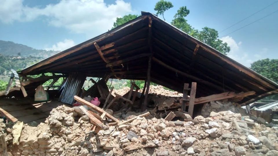 A house whose stone walls have collapsed completely, leaving its heavy timber roof resting flat on a mound of rubble, with hills behind.