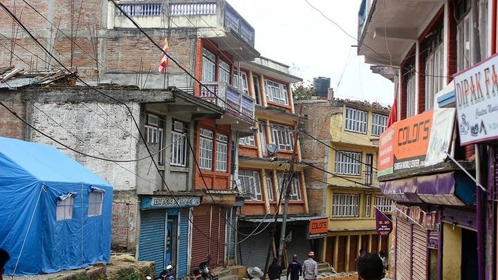 A street of tilted and cracked multistorey buildings with their shutters closed, a blue relief tent pitched on open ground at the left and a few people walking through.