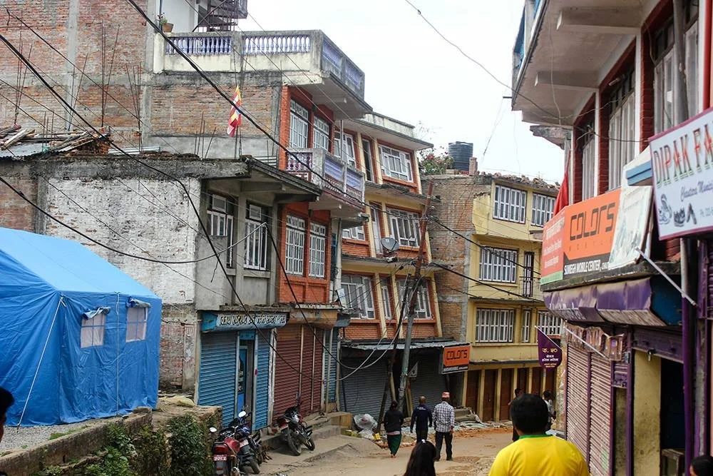 A street of tilted and cracked multistorey buildings with their shutters closed, a blue relief tent pitched on open ground at the left and a few people walking through.