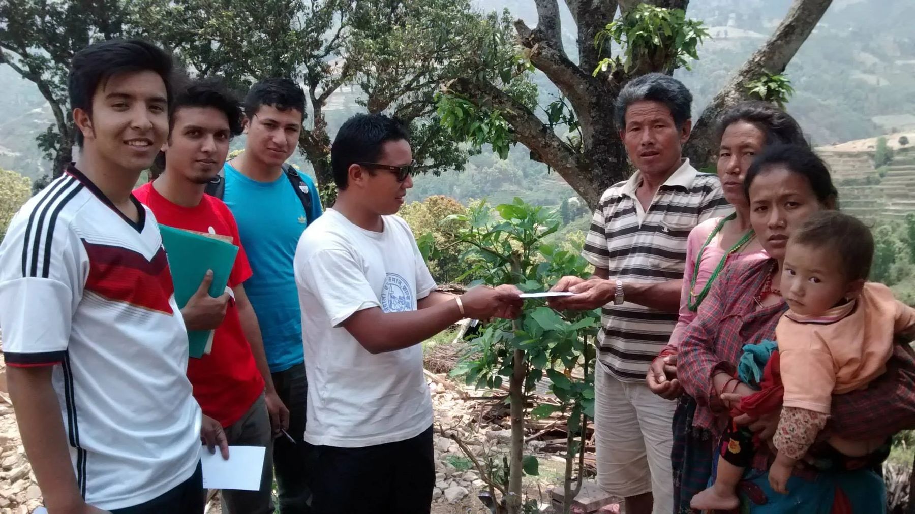 Student volunteers hand an envelope of cash relief to a family standing under a tree on a terraced hillside, with two women and a small child beside them.
