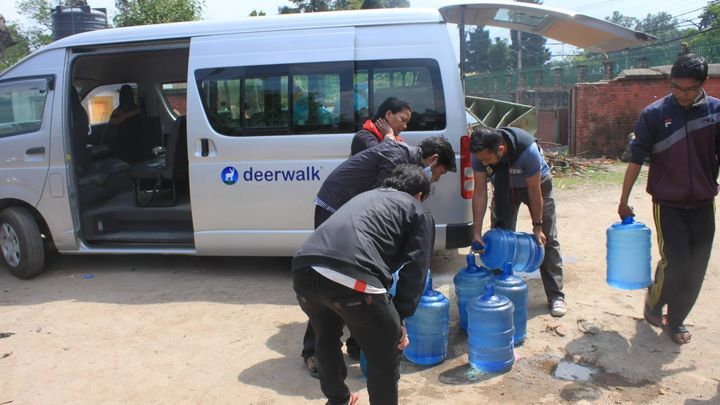 Volunteers lift large blue water jars out of the back of a white van marked deerwalk, lining them up on the ground while another volunteer carries one away.