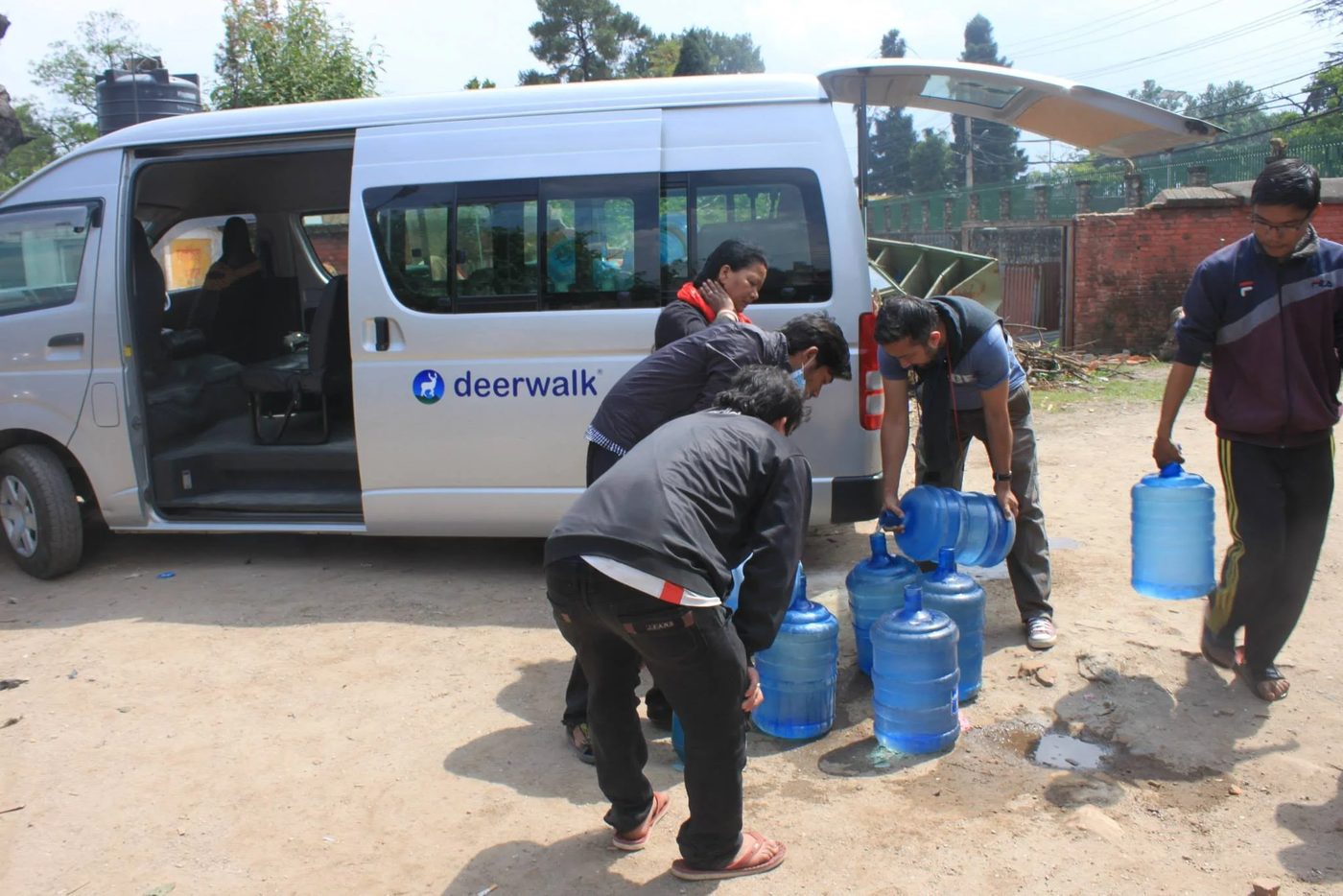 Volunteers lift large blue water jars out of the back of a white van marked deerwalk, lining them up on the ground while another volunteer carries one away.