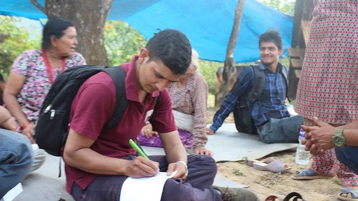 A volunteer sits cross legged on a mat under a blue tarpaulin writing on a sheet of paper, with a woman and other residents seated around him.