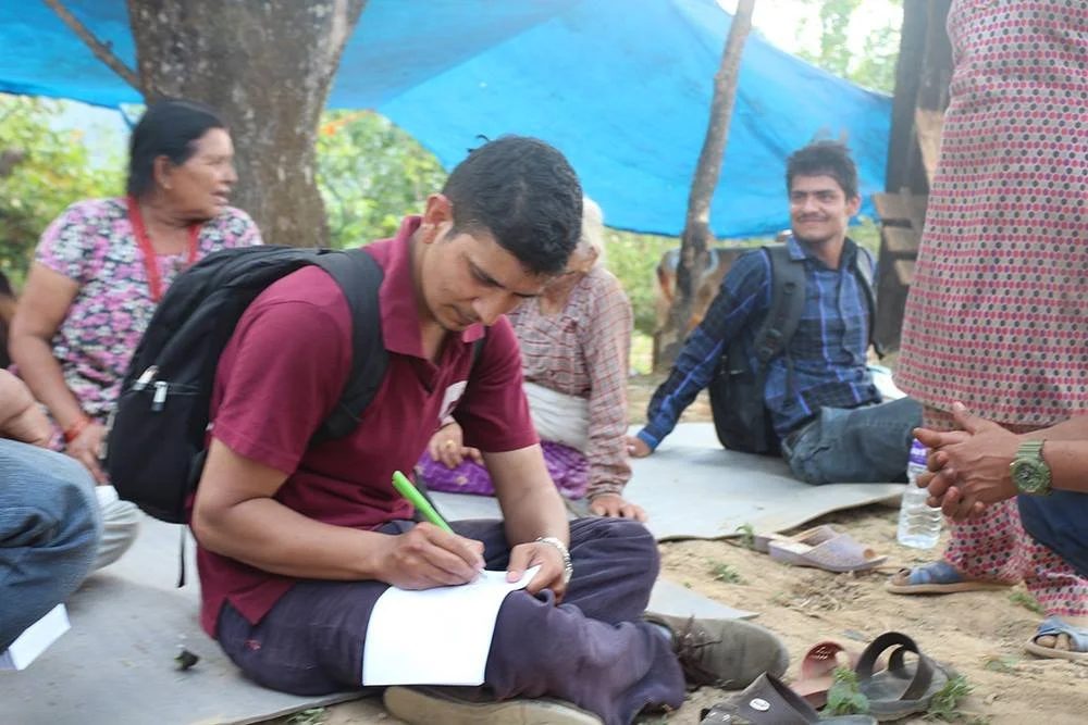 A volunteer sits cross legged on a mat under a blue tarpaulin writing on a sheet of paper, with a woman and other residents seated around him.