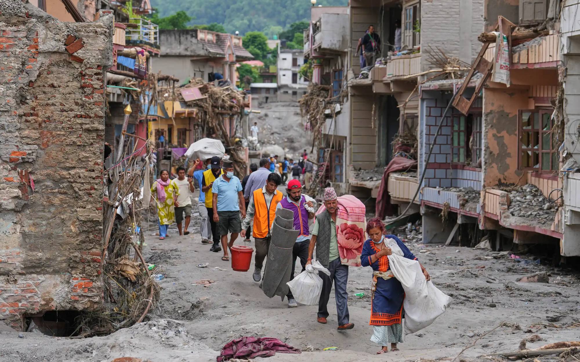 Residents carry salvaged blankets, sacks and belongings along a street buried in grey flood silt, past buildings stripped back to bare concrete.