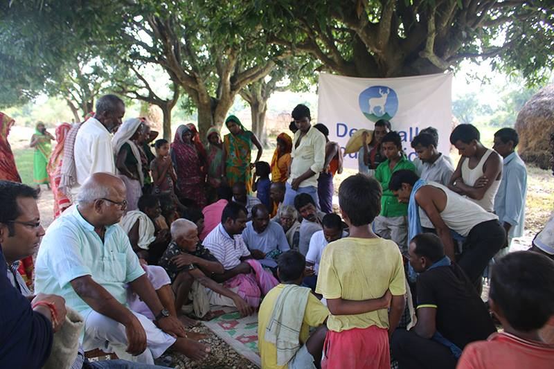 Villagers sit closely together on mats and bare ground under trees, facing a Deerwalk Foundation banner strung between the trunks, with more people standing at the back.