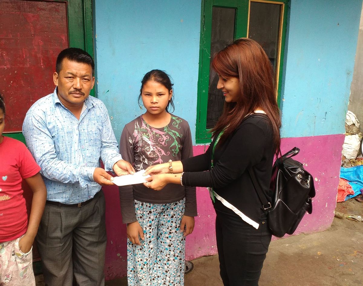 A man and a woman hand an envelope to a girl in a patterned dress standing between them outside the building.
