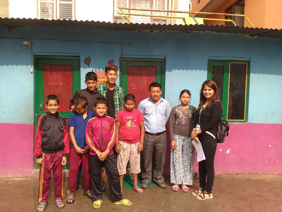 Nine children and three adults stand in a row outside a small building painted pink and turquoise.