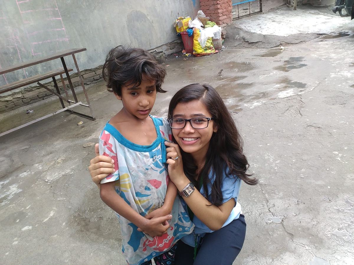A young woman crouches beside a small girl in a blue and white dress on a concrete yard, an arm around her shoulder.