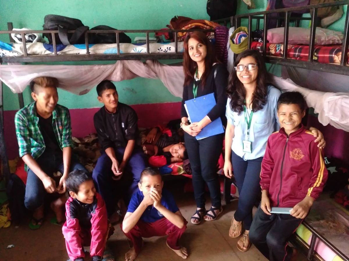 Two visitors stand among six children in front of metal bunk beds stacked with bedding and clothes.