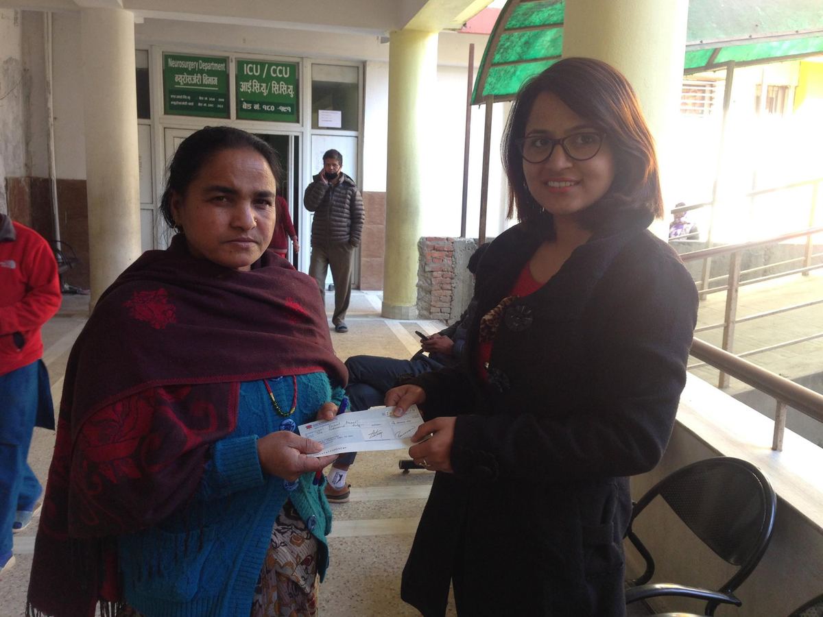 A woman in a maroon shawl takes an envelope from a woman in a black jacket in a hospital corridor.