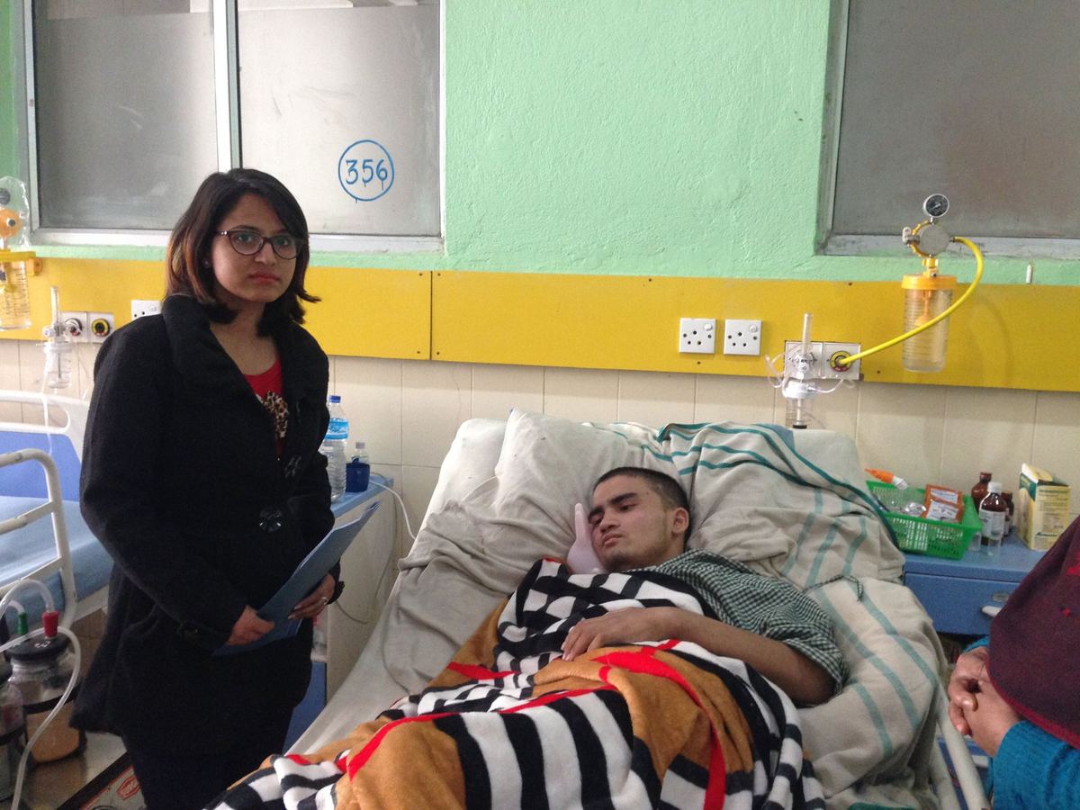 A young man lies in a hospital bed under a striped blanket, an oxygen outlet on the wall behind him, while a woman in a black jacket stands at the bedside.