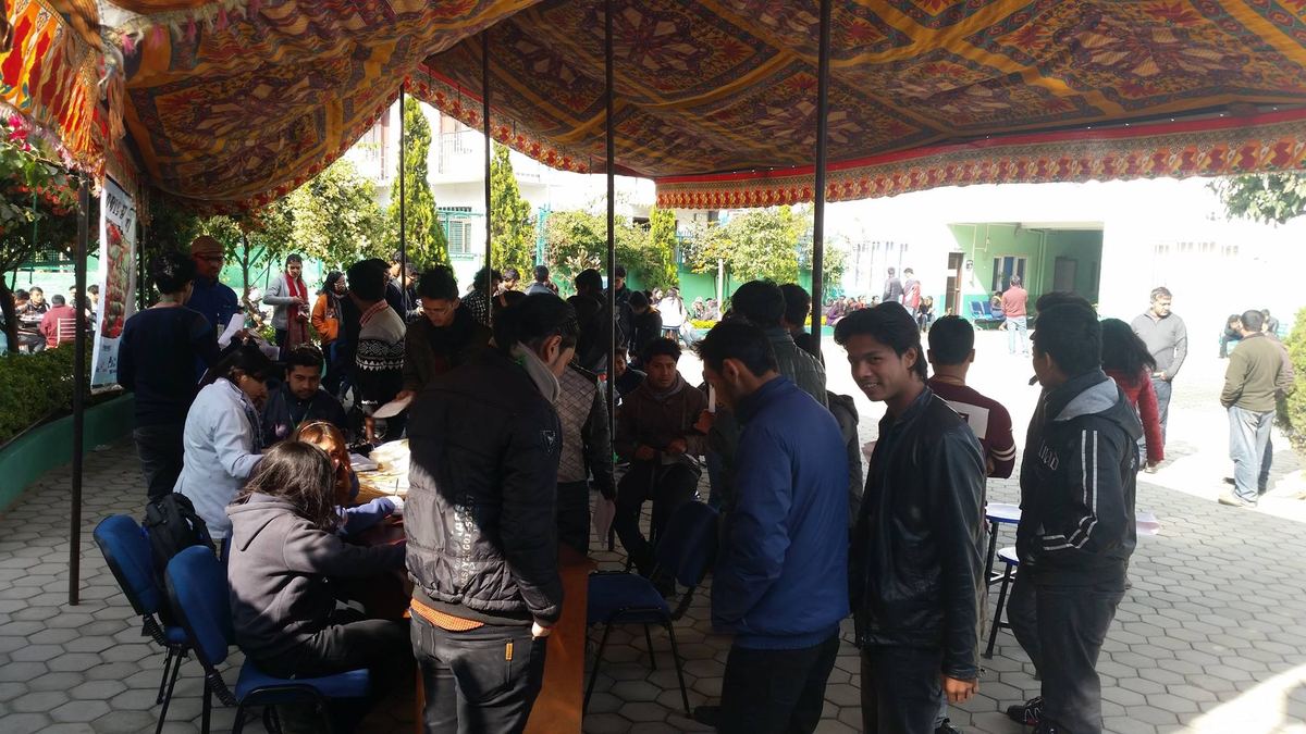A crowd of students queues at tables under a patterned marquee on a paved courtyard.