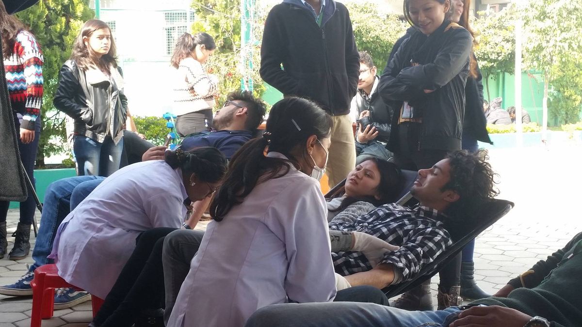 Donors lie on beds outdoors under trees with nurses and onlookers standing over them.