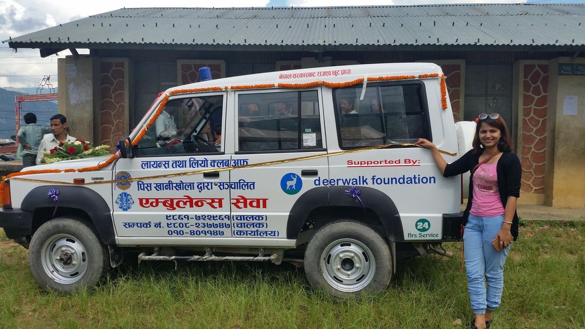 A white Tata Sumo ambulance parked outside a house, lettered in Nepali and carrying a Supported By deerwalk foundation decal on the door, with a woman standing beside it.