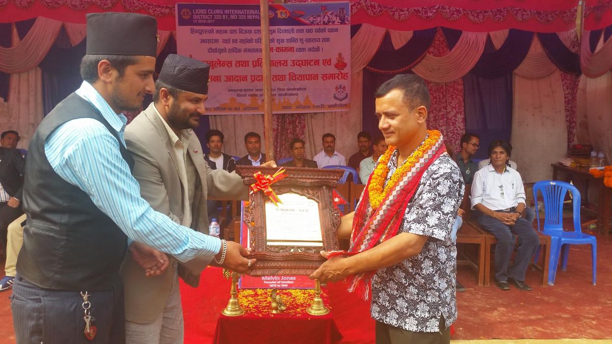 A garlanded man is handed a framed certificate by two men in Dhaka topi on a stage hung with red and pink cloth, with seated guests behind.