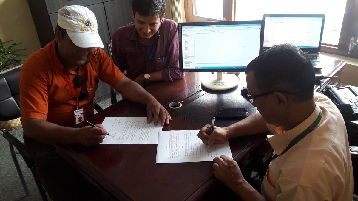 Three men sit around an office desk signing papers, two computer monitors behind them.