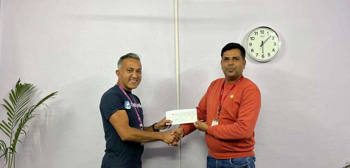 Two men shake hands in an office while holding a cheque between them, with a wall clock and a potted palm behind them.