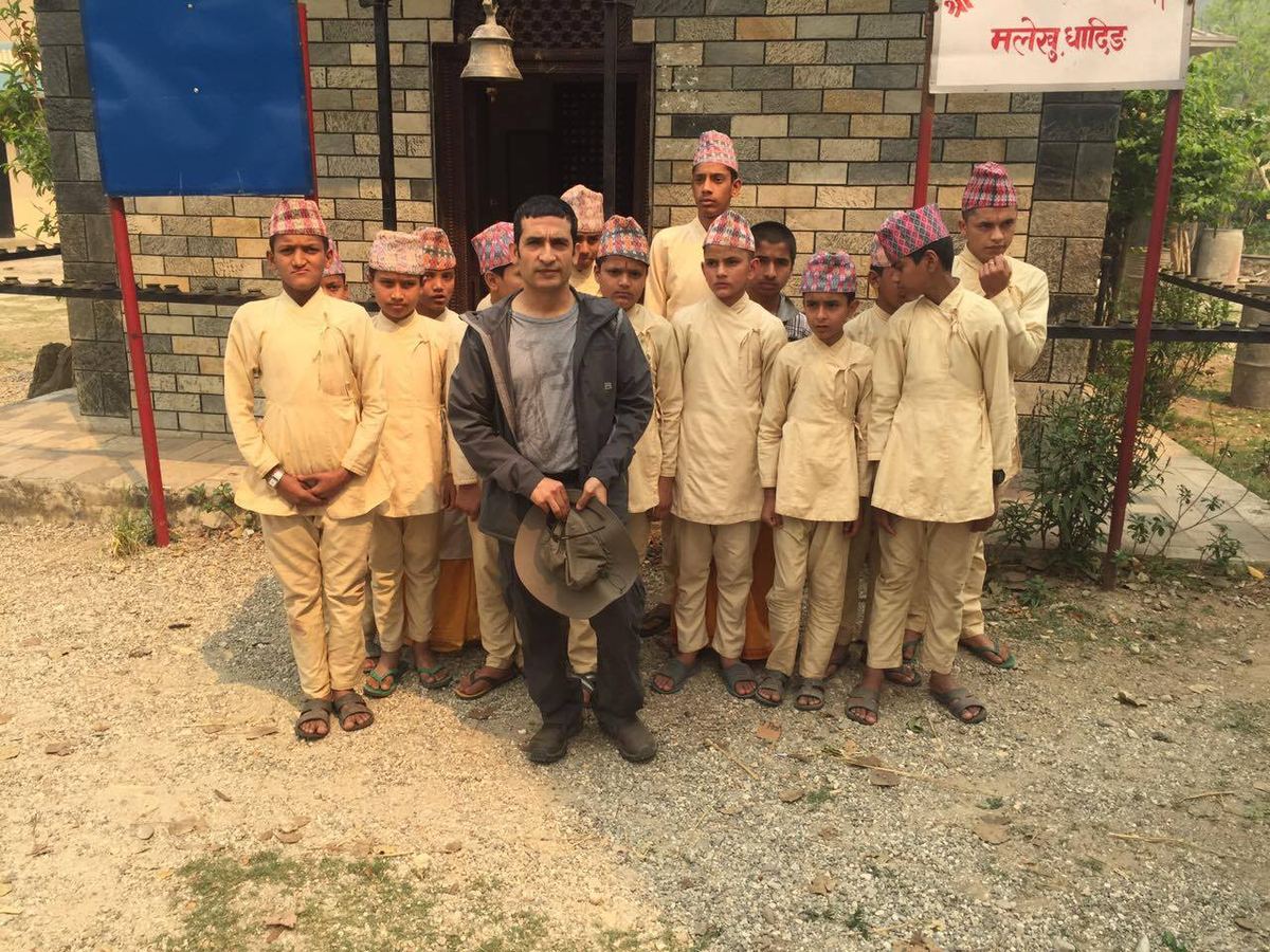 Eleven boys in cream kurta and Dhaka topi stand in a line outside a stone school building with a man in a grey jacket among them.