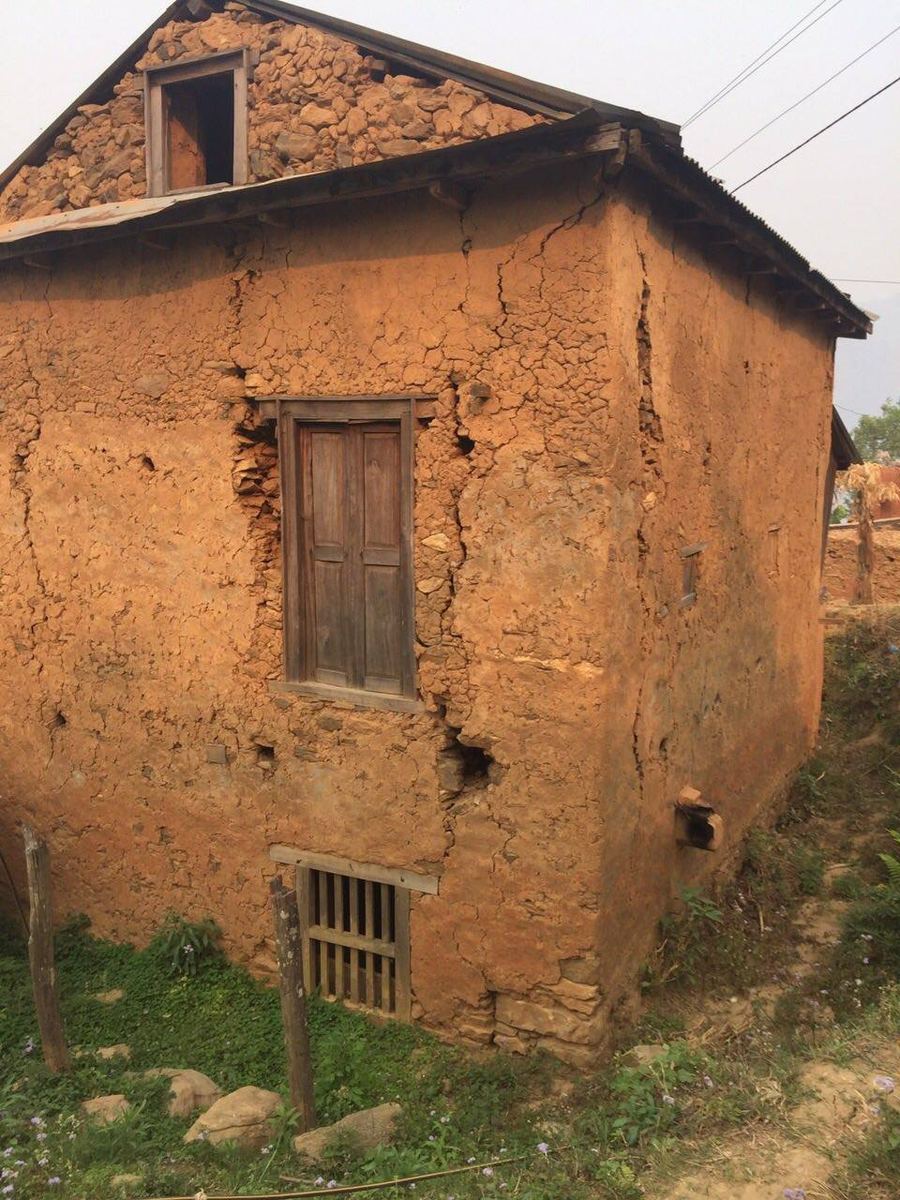 A small mud walled house with a timber door and a cracked upper wall, standing on a grass bank.